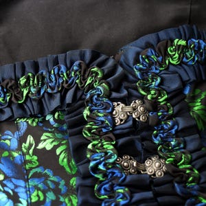 K&ouml;nnte beinhalten: Nahaufnahme eines dunkelblauen Kleidungsst&uuml;cks mit blauen und gr&uuml;nen Blumenmustern. Der Ausschnitt ist mit ger&uuml;schtem Stoff in Blau, Gr&uuml;n und Schwarz verziert. Silberfarbene Verschl&uuml;sse sichern das Kleidungsst&uuml;ck.