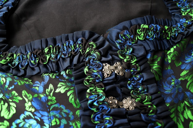 K&ouml;nnte beinhalten: Nahaufnahme eines dunkelblauen Kleidungsst&uuml;cks mit blauen und gr&uuml;nen Blumenmustern. Der Ausschnitt ist mit ger&uuml;schtem Stoff in Blau, Gr&uuml;n und Schwarz verziert. Silberfarbene Verschl&uuml;sse sichern das Kleidungsst&uuml;ck.