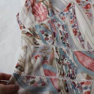 May include: A light-colored, patterned top with a patchwork design. The fabric features a mix of floral and abstract patterns in shades of red, blue, and beige. The top has a tiered construction with delicate lace trim.