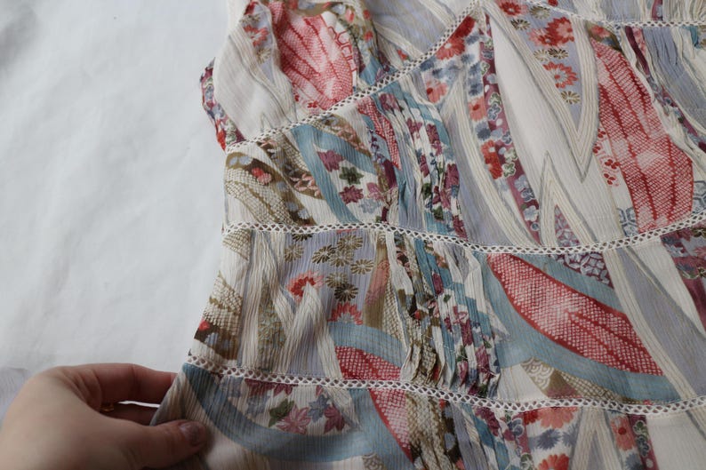 May include: A light-colored, patterned top with a patchwork design. The fabric features a mix of floral and abstract patterns in shades of red, blue, and beige. The top has a tiered construction with delicate lace trim.