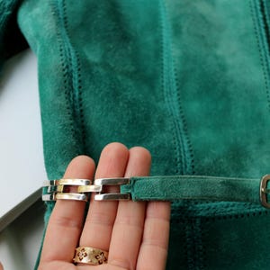 May include: Close-up of a teal suede jacket with a belt and silver and gold buckle. The jacket has vertical stitched panels and a matching belt with a rectangular buckle.