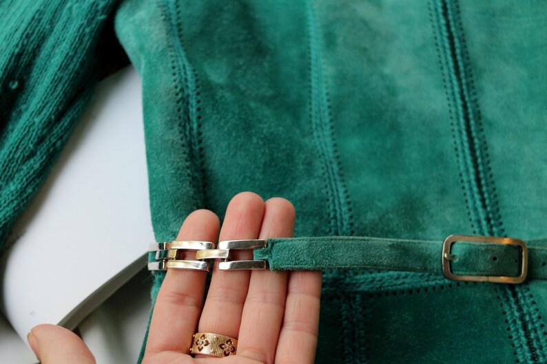 May include: Close-up of a teal suede jacket with a belt and silver and gold buckle. The jacket has vertical stitched panels and a matching belt with a rectangular buckle.