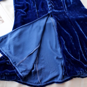 May include: A close-up of a royal blue velvet dress with a high slit. The dress has a button-up front and a flowing skirt. The lining is a lighter shade of blue. The fabric has a soft, luxurious texture.