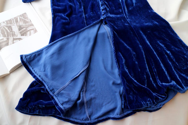 May include: A close-up of a royal blue velvet dress with a high slit. The dress has a button-up front and a flowing skirt. The lining is a lighter shade of blue. The fabric has a soft, luxurious texture.