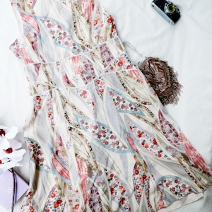May include: A sleeveless, floral-patterned dress in shades of white, pink, red, and blue. The dress features a delicate, flowing design with a V-neckline and a subtle, decorative trim. A small, vintage camera and a fringed purse are also visible.