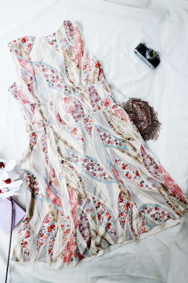 May include: A sleeveless, floral-patterned dress in shades of white, pink, red, and blue. The dress features a delicate, flowing design with a V-neckline and a subtle, decorative trim. A small, vintage camera and a fringed purse are also visible.