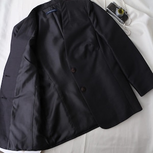May include: A black blazer with a notched collar and two brown buttons. The jacket is open, revealing a dark lining. A vintage camera and a string of pearls are on a white surface.