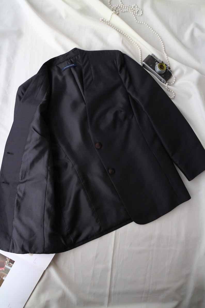 May include: A black blazer with a notched collar and two brown buttons. The jacket is open, revealing a dark lining. A vintage camera and a string of pearls are on a white surface.