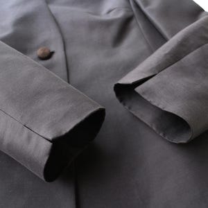 May include: Close-up of a dark gray blazer with a single brown button. The sleeves are visible, with a split cuff design. The fabric has a subtle sheen, suggesting a formal or semi-formal garment.