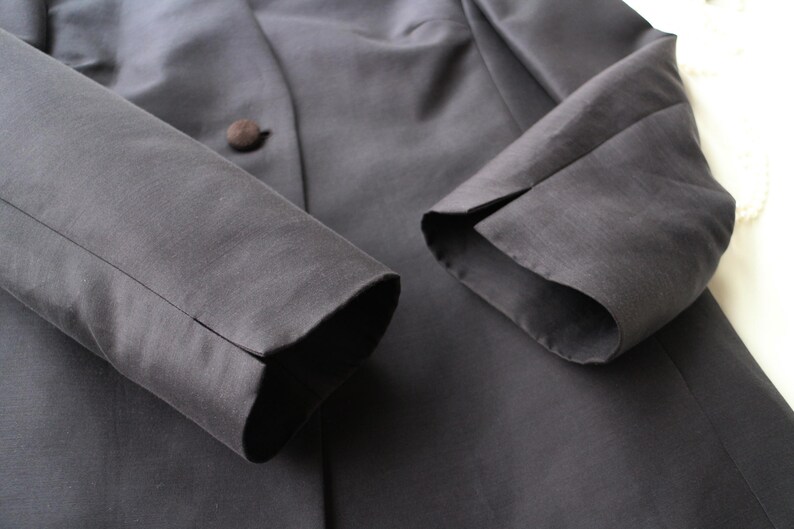 May include: Close-up of a dark gray blazer with a single brown button. The sleeves are visible, with a split cuff design. The fabric has a subtle sheen, suggesting a formal or semi-formal garment.