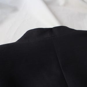 May include: Close-up of a black garment, possibly a jacket or coat, with a visible collar and shoulder seam. The fabric appears smooth and dark, contrasting with the white background. The image focuses on the upper portion of the garment.