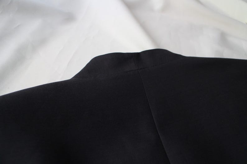 May include: Close-up of a black garment, possibly a jacket or coat, with a visible collar and shoulder seam. The fabric appears smooth and dark, contrasting with the white background. The image focuses on the upper portion of the garment.