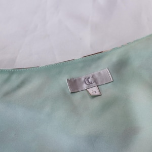 May include: Close-up of a light green satin fabric with a label that reads "CC" and the number "12". The fabric has a subtle sheen and appears to be part of a garment. The background is a plain white.