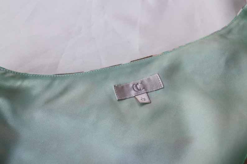 May include: Close-up of a light green satin fabric with a label that reads "CC" and the number "12". The fabric has a subtle sheen and appears to be part of a garment. The background is a plain white.