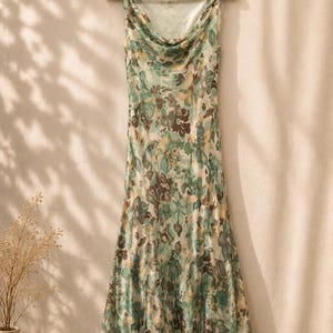May include: A sleeveless, floral-print dress in shades of green, brown, and beige hangs on a wooden hanger. The dress features a cowl neckline and a flowing, midi-length skirt. The floral pattern is repeated throughout the dress.