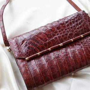 May include: A reddish-brown leather handbag with a textured, crocodile-like pattern. The bag features a long, thin strap and a gold-colored bar with small gold accents. The bag is rectangular and has a flap closure.