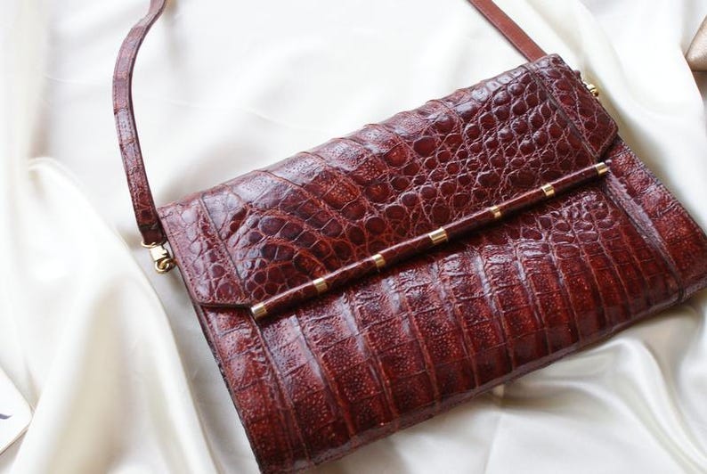 May include: A reddish-brown leather handbag with a textured, crocodile-like pattern. The bag features a long, thin strap and a gold-colored bar with small gold accents. The bag is rectangular and has a flap closure.