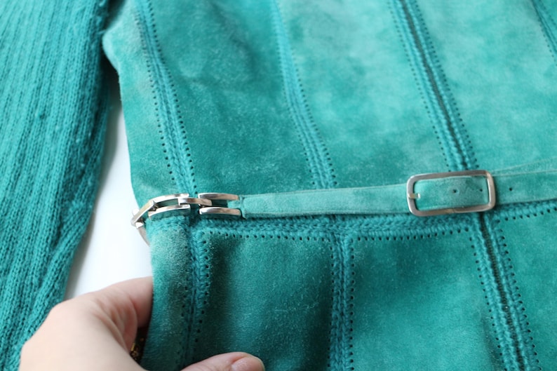 May include: Teal suede jacket with a matching belt and silver buckle. The jacket features vertical stitched panels and a knitted teal sleeve. The belt is fastened with a silver buckle. The jacket is a vintage style.