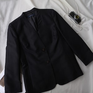 May include: A black blazer with a notched collar and two-button closure. The long-sleeved jacket is made of a smooth fabric and is displayed on a white surface. A vintage camera and a string of pearls are also visible.