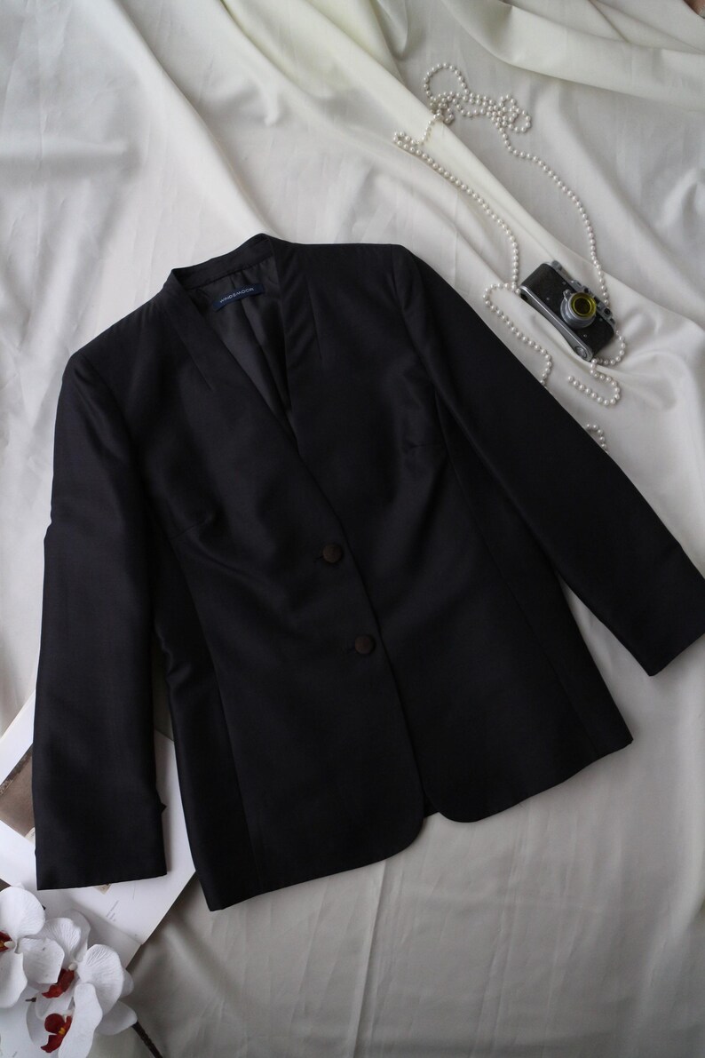 May include: A black blazer with a notched collar and two-button closure. The long-sleeved jacket is made of a smooth fabric and is displayed on a white surface. A vintage camera and a string of pearls are also visible.