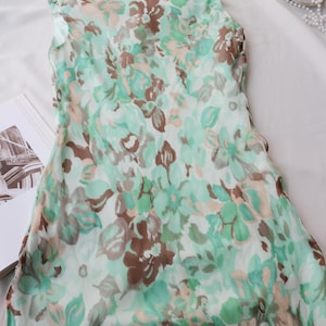 May include: A sleeveless dress with a floral pattern in shades of green, brown, and peach. The dress is made of a lightweight fabric and is lying on a white surface. The floral print is detailed and covers the entire dress.