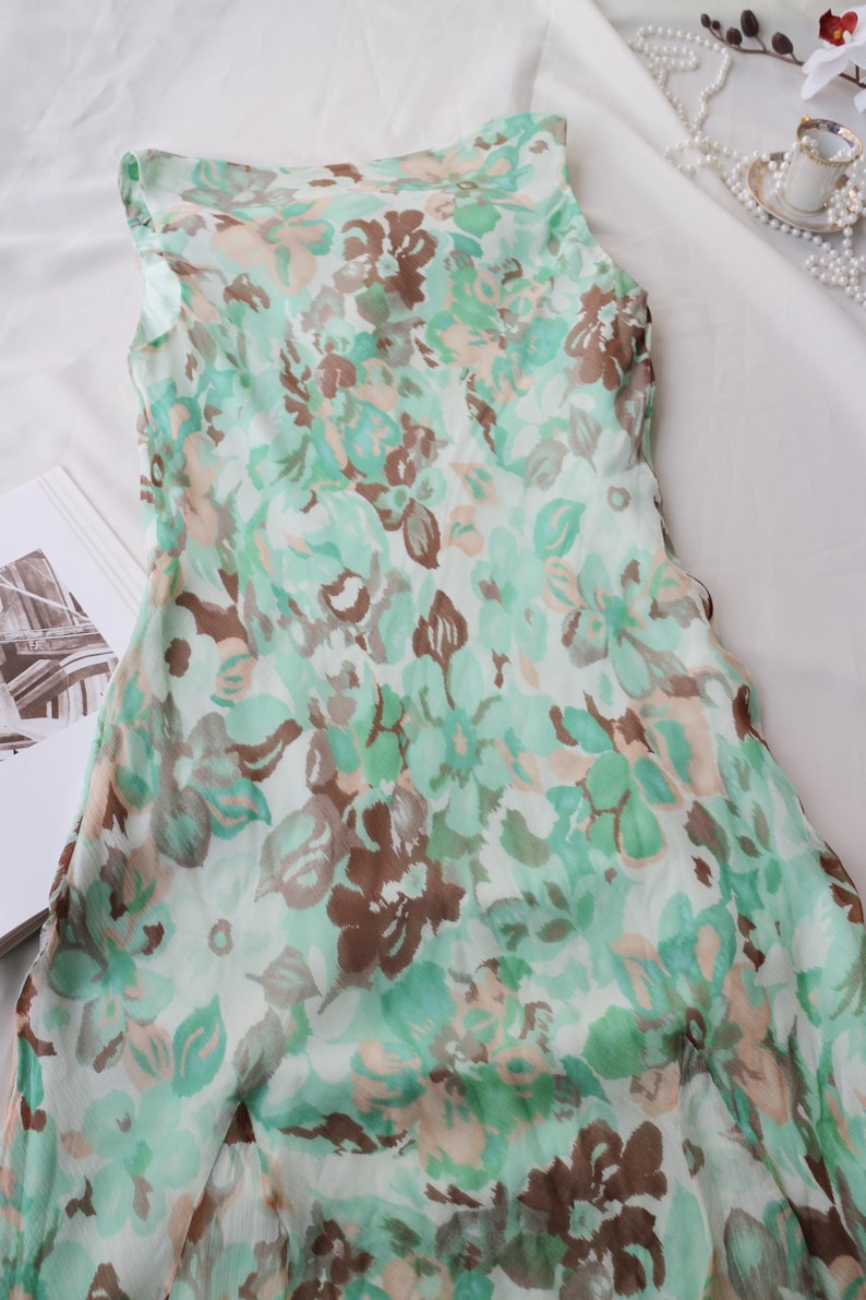 May include: A sleeveless dress with a floral pattern in shades of green, brown, and peach. The dress is made of a lightweight fabric and is lying on a white surface. The floral print is detailed and covers the entire dress.