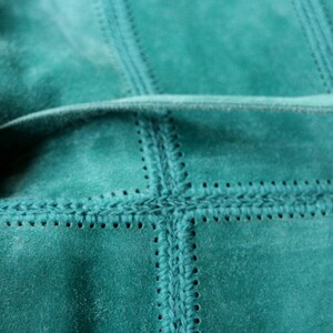 May include: Close-up of a teal suede fabric with a stitched, grid-like pattern. The stitching creates a textured effect, with visible seams and decorative details. The fabric is soft and appears to be high-quality.