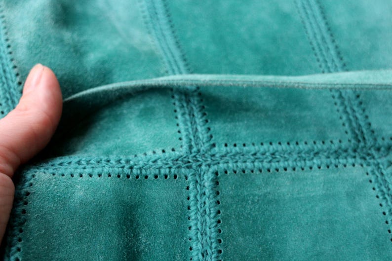 May include: Close-up of a teal suede fabric with a stitched, grid-like pattern. The stitching creates a textured effect, with visible seams and decorative details. The fabric is soft and appears to be high-quality.