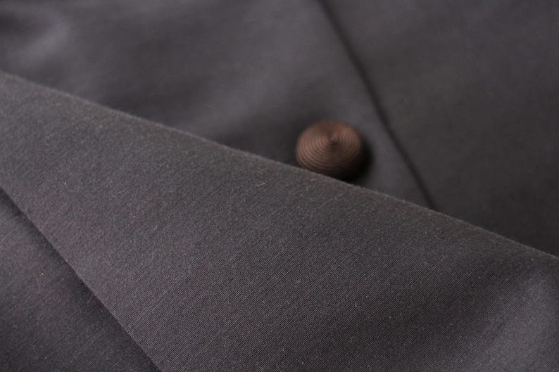 May include: Close-up of dark gray fabric with a brown button. The fabric appears to be a woven material, possibly part of a garment. The button is round and has a textured surface.
