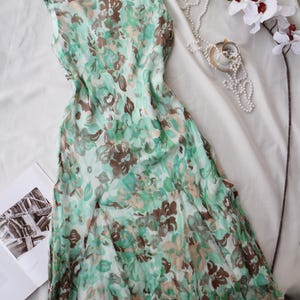 May include: A sleeveless, flowing dress with a floral pattern in shades of green, brown, and beige. The dress is draped on a white surface, with a pearl necklace, a teacup, and a sprig of white orchids nearby. The dress appears to be made of a lightweight fabric.