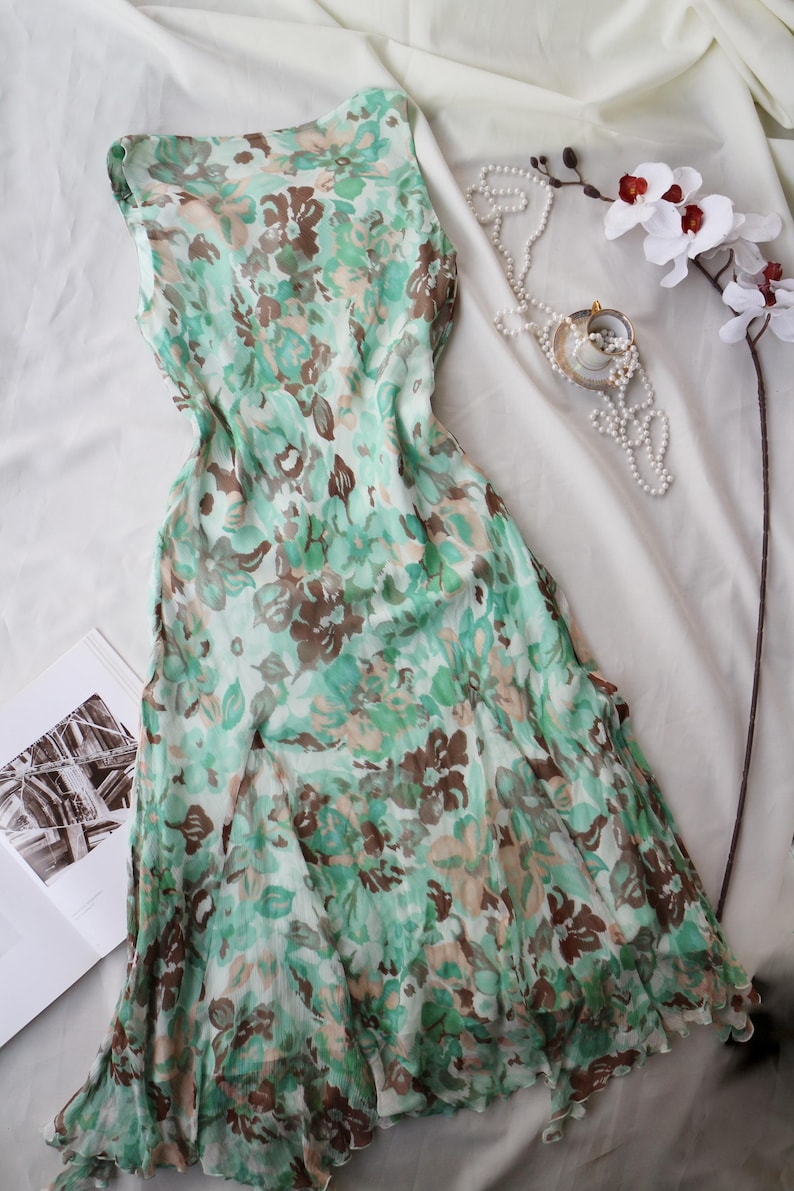 May include: A sleeveless, flowing dress with a floral pattern in shades of green, brown, and beige. The dress is draped on a white surface, with a pearl necklace, a teacup, and a sprig of white orchids nearby. The dress appears to be made of a lightweight fabric.