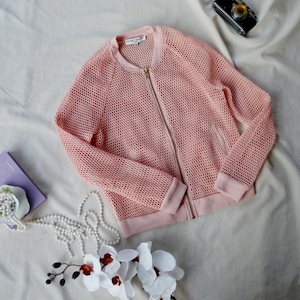 May include: A pale pink mesh bomber jacket with a gold zip, cuffs, and a collar. The jacket is displayed on a cream-coloured surface, with a vintage camera, a string of pearls, and white orchids nearby.