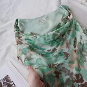 May include: A flowing, floral-print dress in shades of green, brown, and white. The dress features a draped neckline and a lightweight, semi-sheer fabric. The dress is folded, revealing the inner lining.