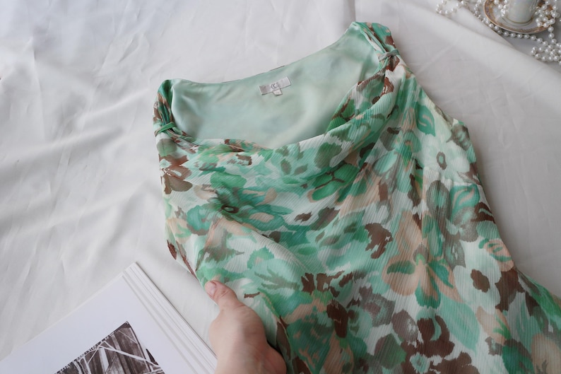 May include: A flowing, floral-print dress in shades of green, brown, and white. The dress features a draped neckline and a lightweight, semi-sheer fabric. The dress is folded, revealing the inner lining.