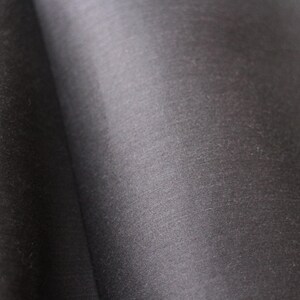 May include: Close-up of a dark gray fabric with a subtle sheen. The material appears to be smooth and possibly woven, with gentle folds creating shadows and highlights. The overall texture suggests a luxurious textile.