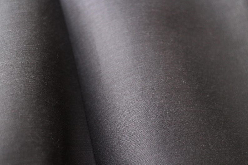May include: Close-up of a dark gray fabric with a subtle sheen. The material appears to be smooth and possibly woven, with gentle folds creating shadows and highlights. The overall texture suggests a luxurious textile.