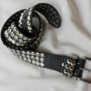 Pode incluir: Um cinto de couro preto com três filas de tachas metálicas prateadas ao longo da tira. O cinto inclui uma fivela de metal prateado e um passador a condizer, com tachas adicionais a decorar a zona perto da fivela.