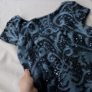 May include: A vintage blue dress with short sleeves and a scoop neckline. The dress is covered in intricate black and silver sequin detailing, creating a swirling pattern. The fabric appears soft and drapes gently. The dress is a classic piece.