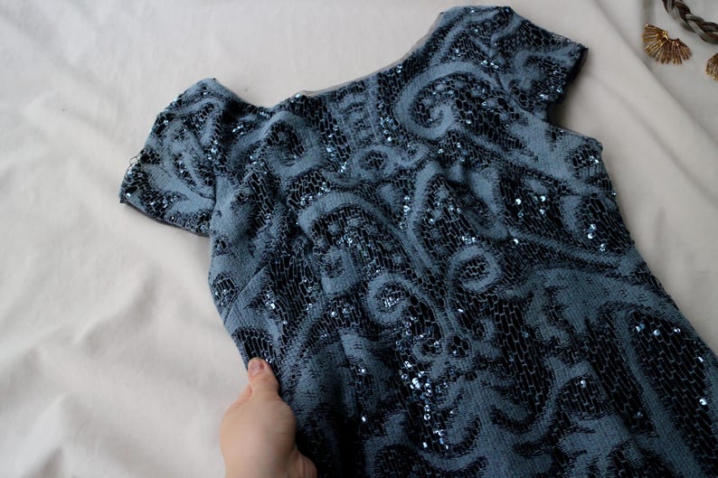 May include: A vintage blue dress with short sleeves and a scoop neckline. The dress is covered in intricate black and silver sequin detailing, creating a swirling pattern. The fabric appears soft and drapes gently. The dress is a classic piece.