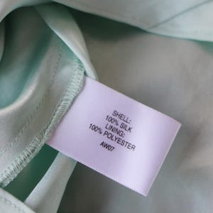 May include: Close-up of a light green silk fabric with a label attached. The label reads "SHELL: 100% SILK, LINING: 100% POLYESTER, AW07". The fabric has a smooth, shiny texture.
