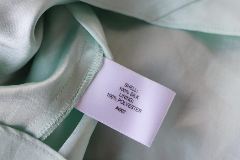 May include: Close-up of a light green silk fabric with a label attached. The label reads "SHELL: 100% SILK, LINING: 100% POLYESTER, AW07". The fabric has a smooth, shiny texture.