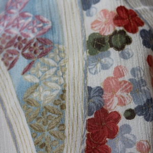 May include: Close-up of a fabric with a floral pattern. The design features flowers in shades of red, pink, and blue, set against a light background with vertical stripes. The fabric has a textured appearance, suggesting a soft material.