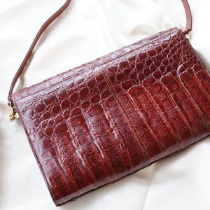 May include: A rectangular, brown leather clutch with a textured, crocodile-like pattern. The bag has a thin, brown strap and gold-colored hardware. The clutch is set against a white, silky background.