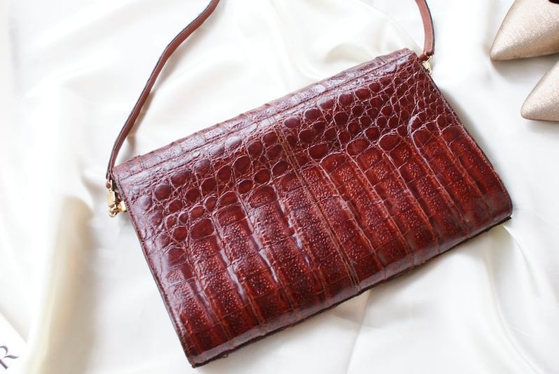 May include: A rectangular, brown leather clutch with a textured, crocodile-like pattern. The bag has a thin, brown strap and gold-colored hardware. The clutch is set against a white, silky background.