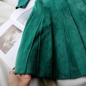 May include: A close-up of a teal green suede skirt with a pleated design. The skirt is laid out on a white surface with a book open in the background. The skirt has a soft, textured appearance.