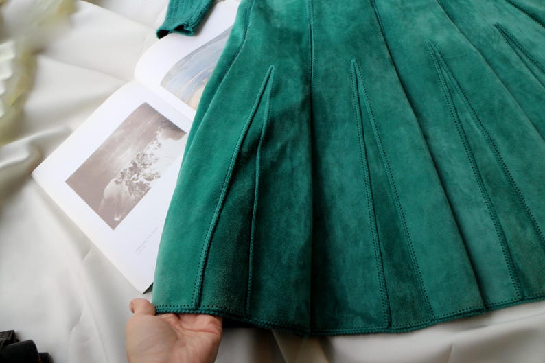 May include: A close-up of a teal green suede skirt with a pleated design. The skirt is laid out on a white surface with a book open in the background. The skirt has a soft, textured appearance.