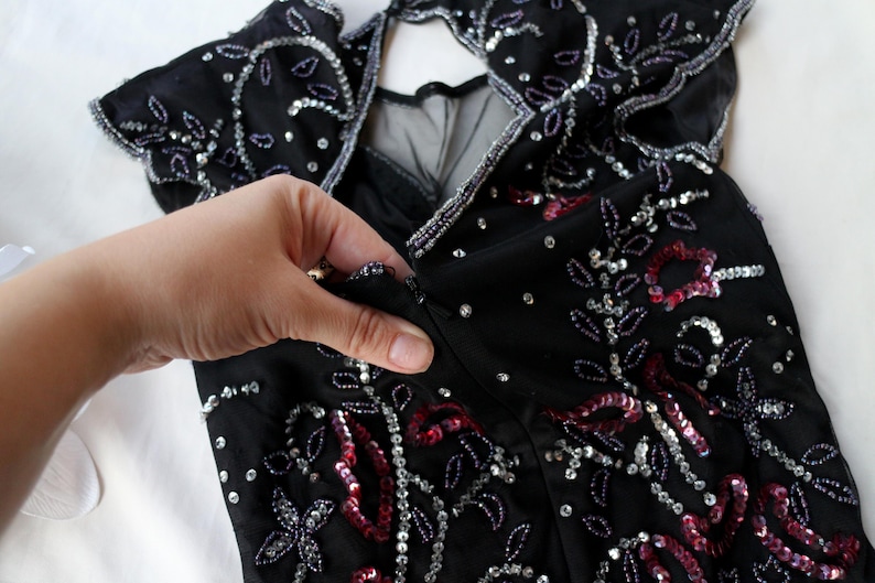 May include: Black dress with silver and red sequin and bead embellishments. The dress features a keyhole back and a zipper closure. The design includes floral and leaf patterns.