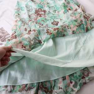 May include: A floral print dress with a light green lining. The outer layer features a pattern of green, brown, and white flowers. The dress is made of a lightweight fabric, ideal for warm weather. The dress is laying on a white surface.