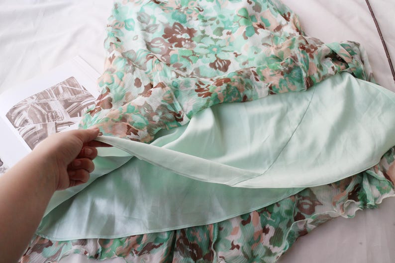 May include: A floral print dress with a light green lining. The outer layer features a pattern of green, brown, and white flowers. The dress is made of a lightweight fabric, ideal for warm weather. The dress is laying on a white surface.