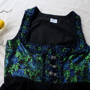 K&ouml;nnte beinhalten: Ein schwarzes Dirndl-Oberteil mit einem Blumenmuster in Blau und Gr&uuml;n. Das Oberteil hat einen quadratischen Ausschnitt und ist mit marineblauen R&uuml;schen verziert. Silberfarbene Verschl&uuml;sse zieren die Vorderseite.